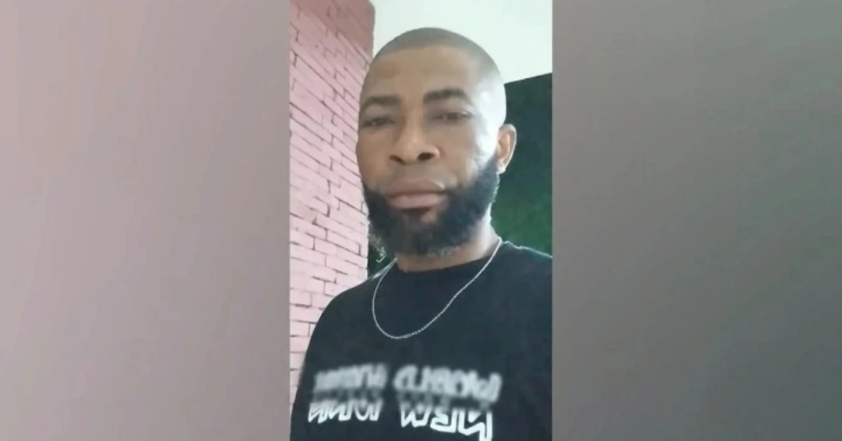 Man with a trimmed beard and short hair, wearing a black T-shirt and a silver chain, standing beside a brick wall.