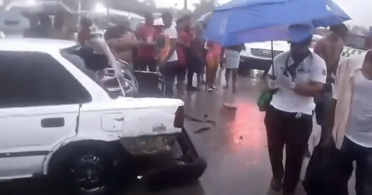 Crowd of people standing in rain around a wrecked white SUV on a wet street, some holding umbrellas.