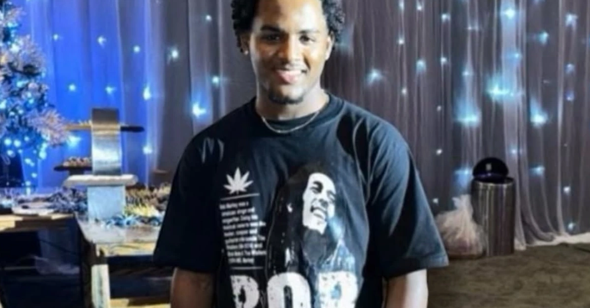 Smiling man in a black graphic t-shirt at a festive event with blue lights and a dessert table behind him.