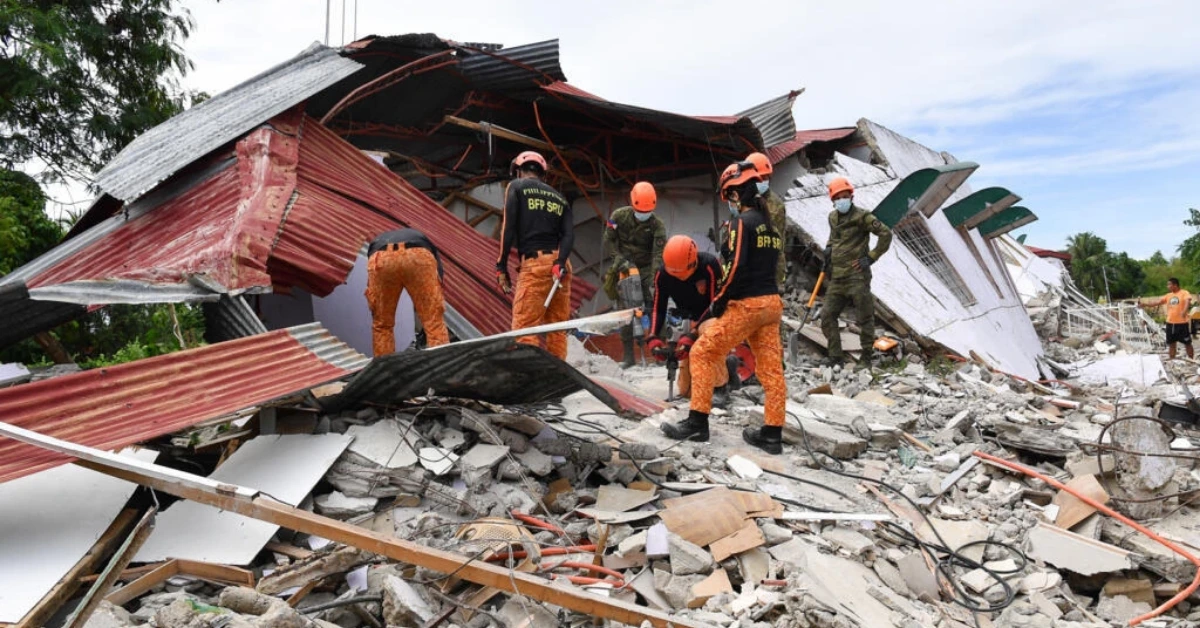 Rescue workers in orange pants and helmets search through rubble of a collapsed building with exposed red metal roofing and debris scattered around.