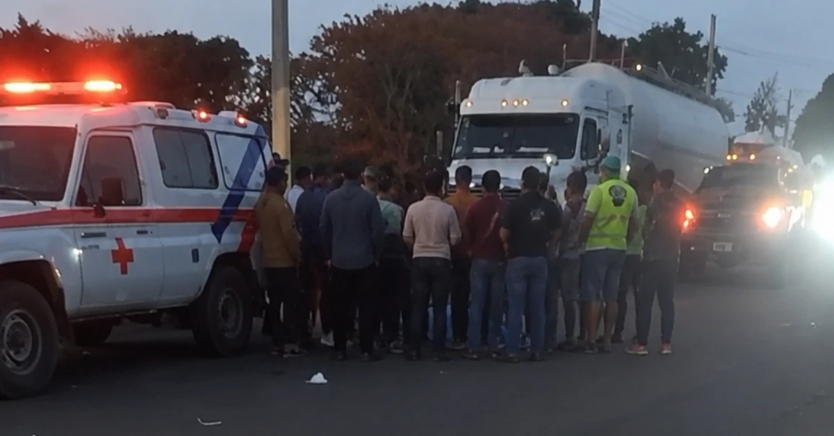 Group of people crowded around a parked emergency vehicle and a large tanker truck at dusk, likely an accident scene.