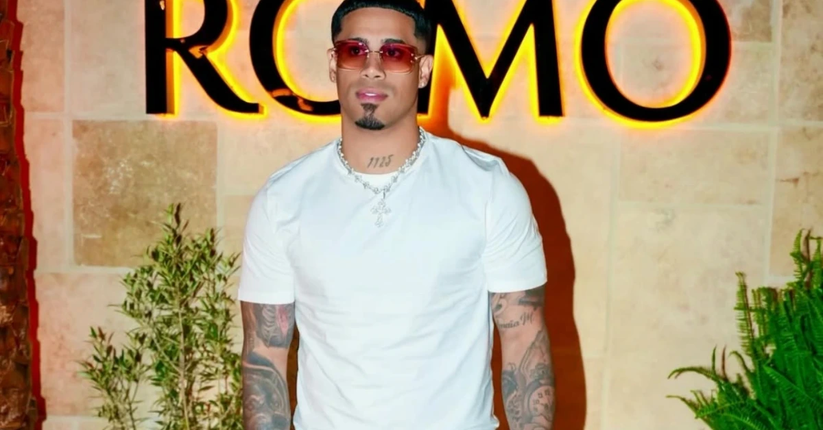 Man in a white T-shirt with tattoos stands in front of a neon ROMO sign, wearing sunglasses and a chain.