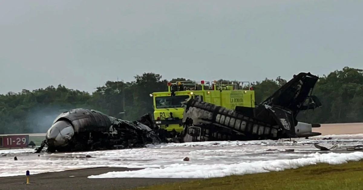 Damaged aircraft fuselage wreckage on a runway with foam and a yellow firefighting truck in the background.