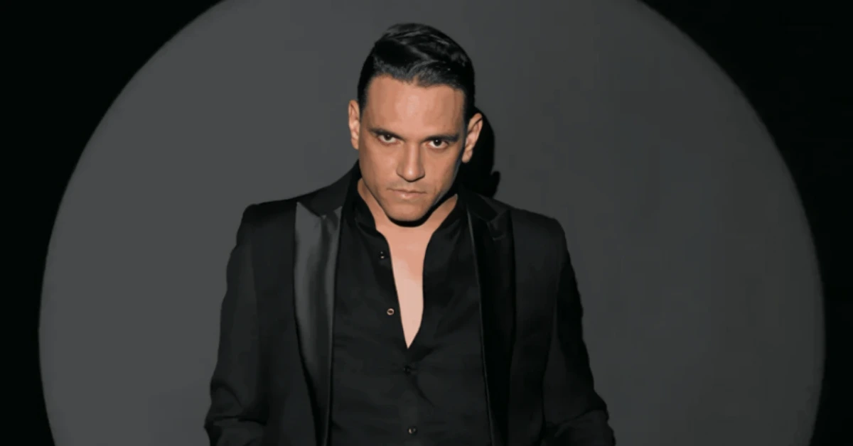 Man in a black suit posing against a dark backdrop with a large gray circle behind him.