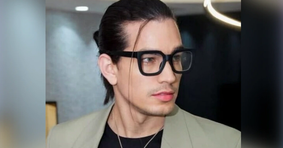 Portrait of a man with slicked-back hair wearing large black square glasses and a light blazer, looking to the right.
