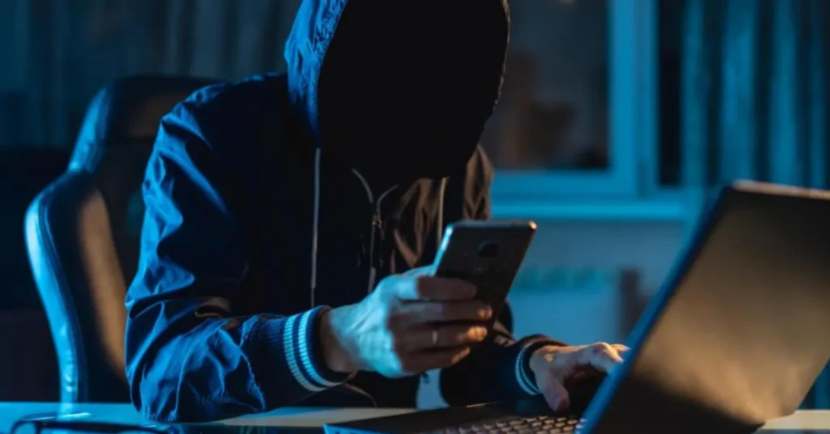 Person wearing a hooded jacket with face obscured, looking at a smartphone while a laptop sits on the desk in a dimly lit blue room.