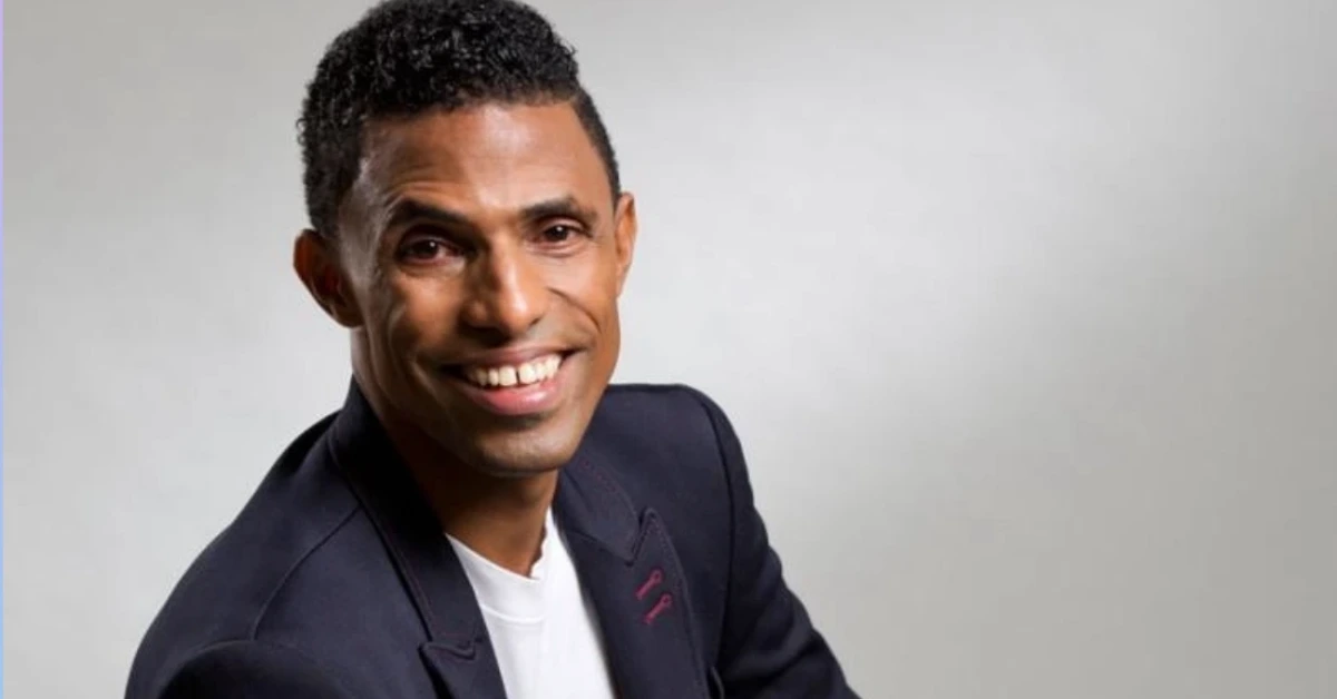 Smiling man in a navy blazer and white shirt posed for a professional headshot against a light gray background, showing shoulders and head.