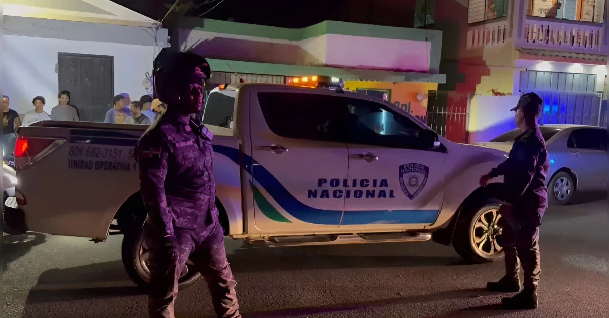 Nighttime street scene with a Policía Nacional pickup and two officers in tactical gear standing near it, colorful buildings in the background and onlookers observing from the sidewalk.
