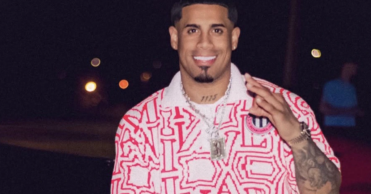 Smiling man with a goatee and tattoos on his forearm, wearing a red and white geometric shirt and necklace at night.