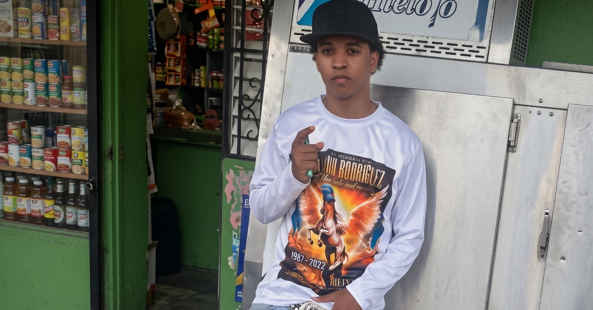 Young man wearing a cap and white graphic long-sleeve shirt points at the camera outside a storefront.