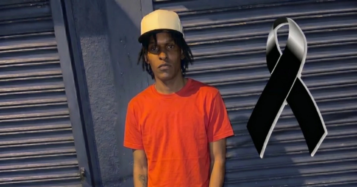 A young person in a red T-shirt and beige cap stands against a blue corrugated wall, with a large black ribbon graphic to the right, signaling mourning or awareness.