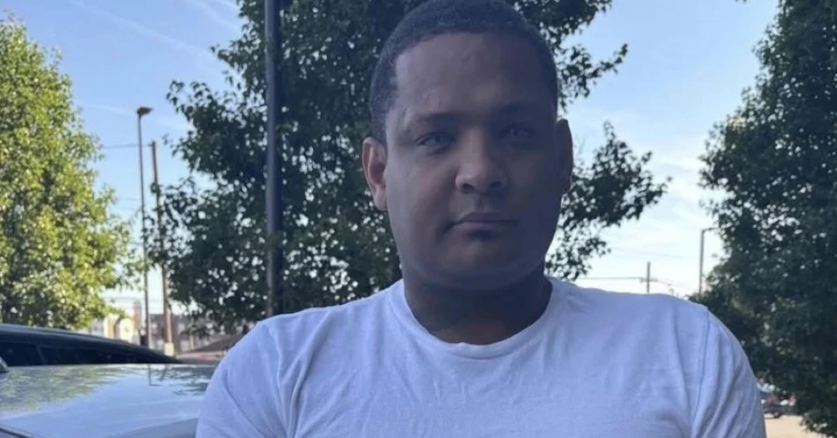 Man in a white T-shirt standing outdoors, looking at the camera with trees and a parked car behind him on a sunny day.