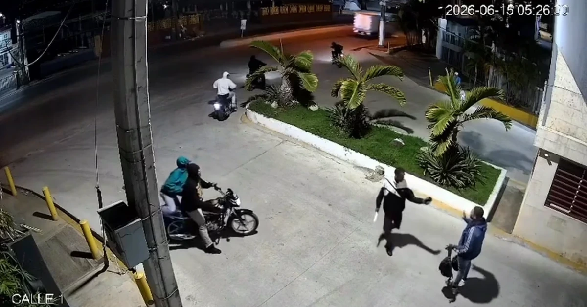 Nighttime street scene with a landscaped median island and palm trees; two people on motorcycles near the foreground and a group walking nearby.