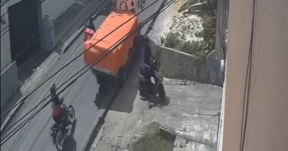 Surveillance image of an orange dump truck blocking a narrow urban street, with a motorbike rider on the left and two people near a wall on the right.
