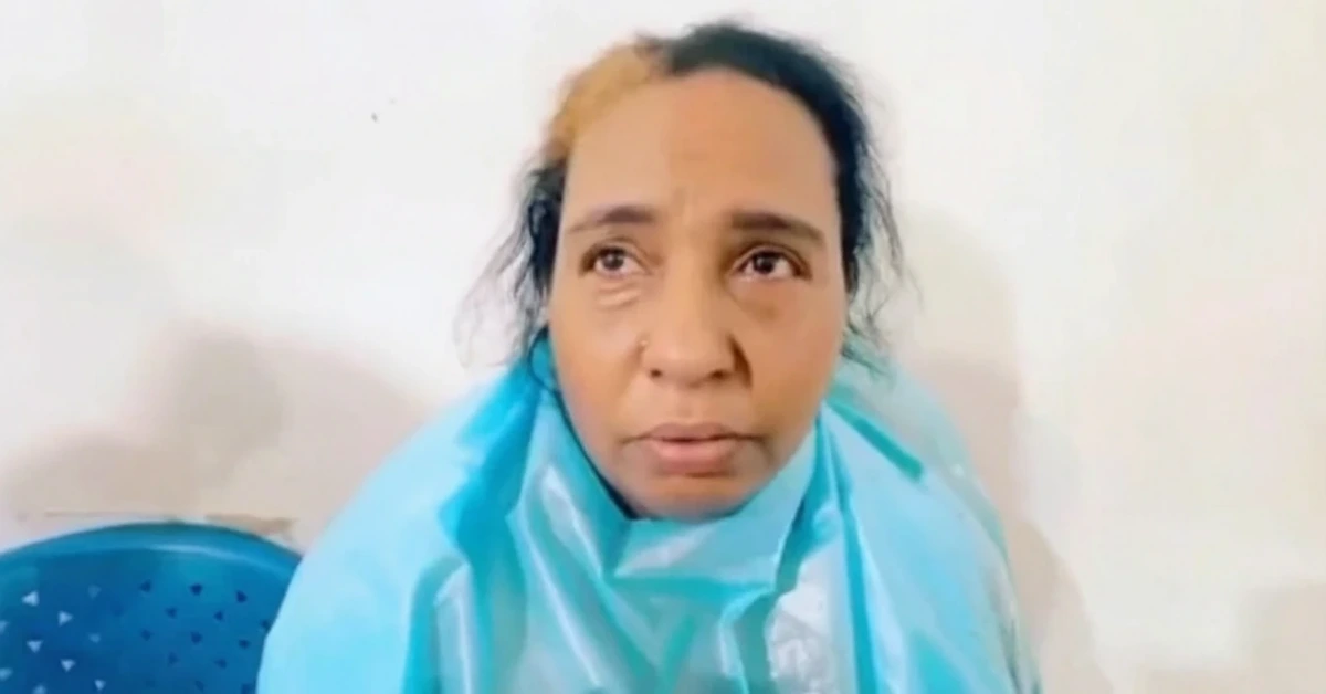 Woman wearing a light blue protective gown seated against a plain wall, looking anxious or worried.