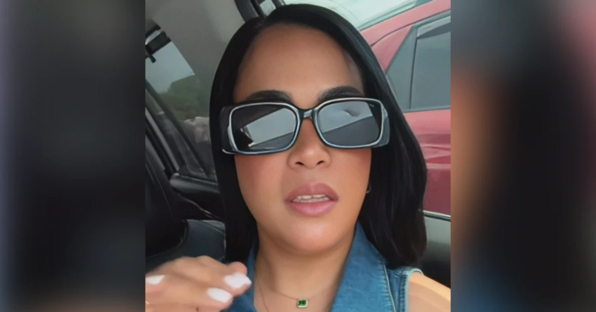 Woman in a car wearing oversized sunglasses and a denim jacket, speaking to the camera with pink lipstick and a green pendant necklace.