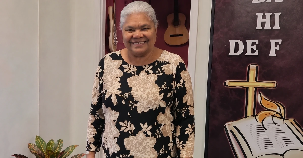 Smiling woman with gray hair in a beige floral dress stands indoors next to a church banner with a cross and Bible.