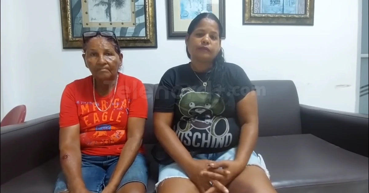Two women sit on a gray sofa in a living room; the older woman wears a red t‑shirt, the younger a black Moschino tee with a teddy bear design on the chest.