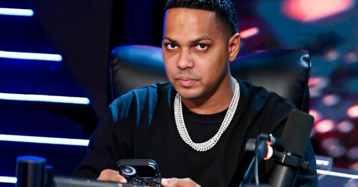 Man in a black shirt with a thick silver chain sits at a studio desk with a microphone, looking at the camera.