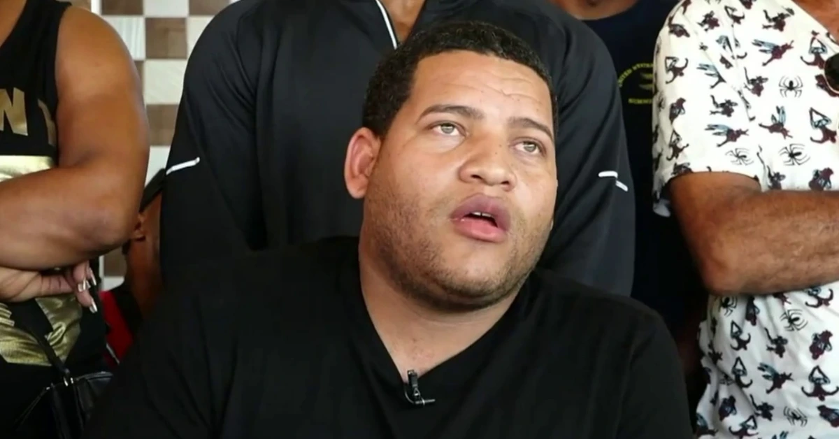 Center image of a man with short hair and stubble wearing a black shirt, looking upward with his mouth slightly open, a clip-on mic on his shirt as people stand around him.