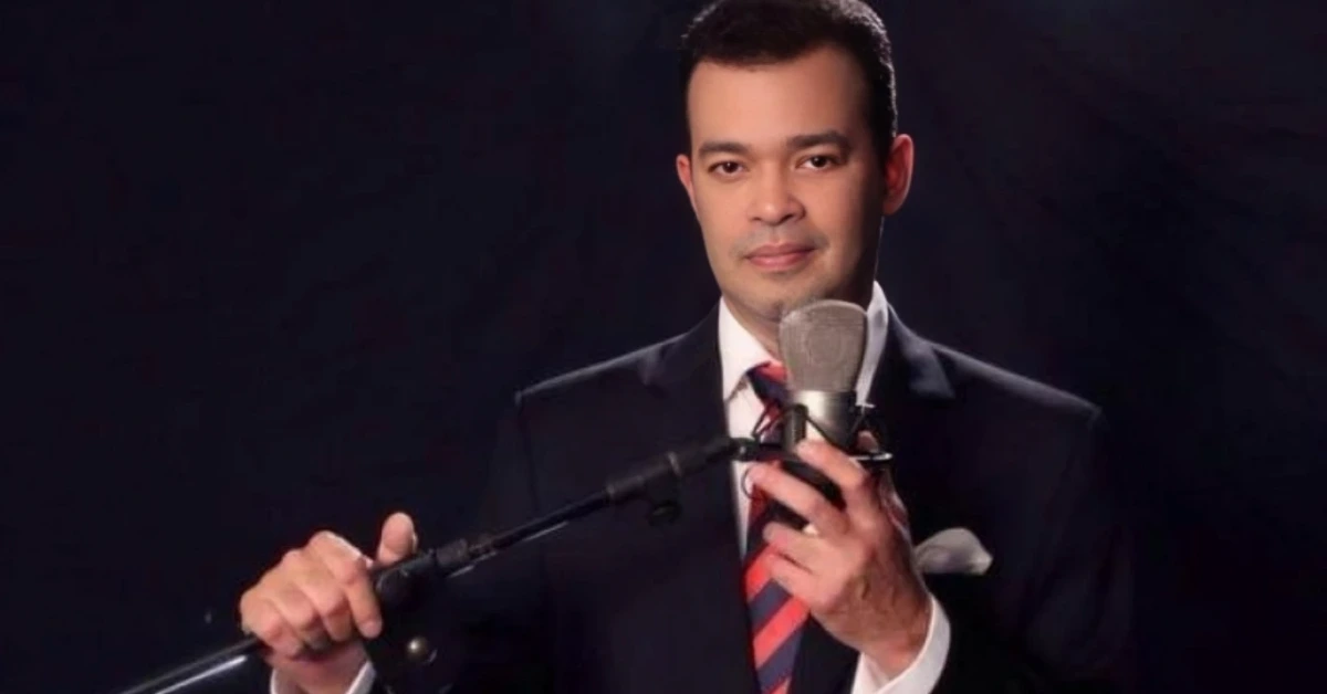 Man in a dark suit and red striped tie speaks into a studio microphone, against a dark background.