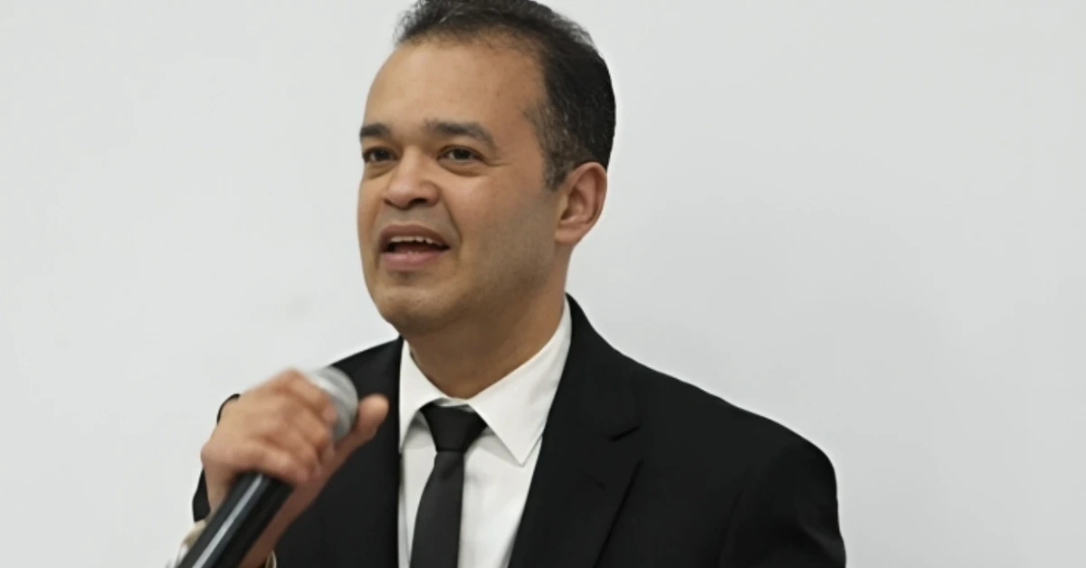 A man in a black suit and tie speaks into a handheld microphone at a formal event.
