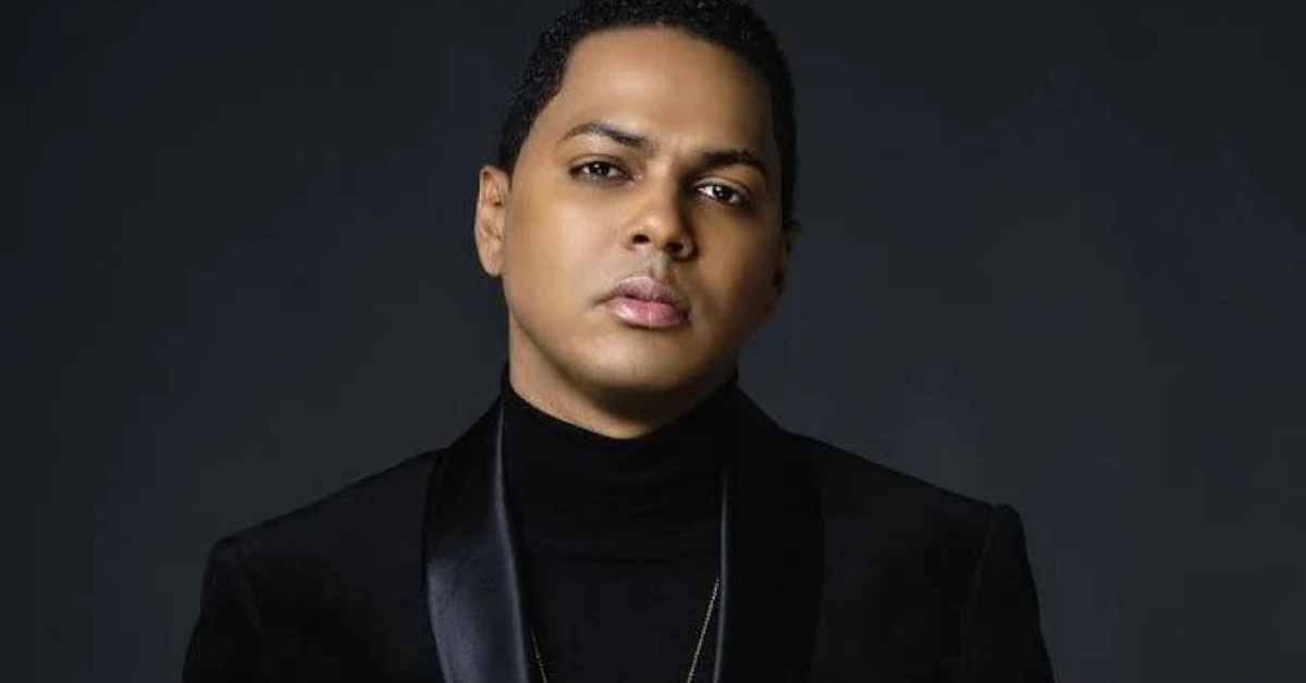 Portrait of a person with short hair wearing a black turtleneck and blazer against a dark gray background.