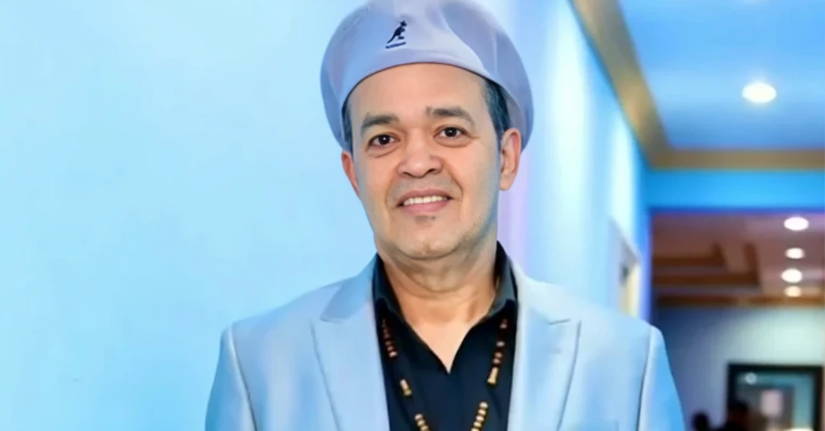 Smiling man in a light-blue blazer and dark shirt, wearing a light-colored cap, standing in a hallway