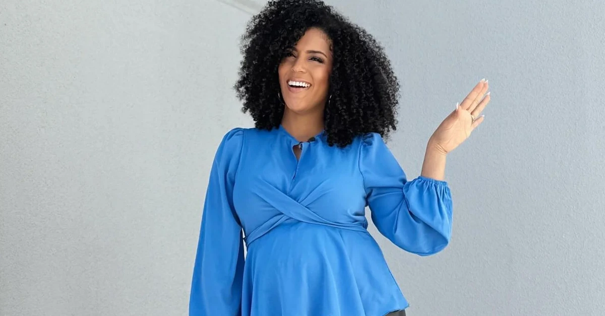 Smiling pregnant woman in a bright blue top waves with her right hand against a light gray wall.