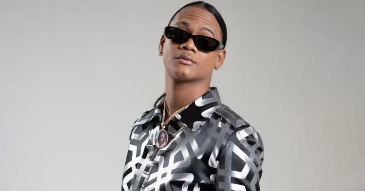 Person posing in black sunglasses with a geometric black-and-white shirt and a round pendant necklace against a light background.