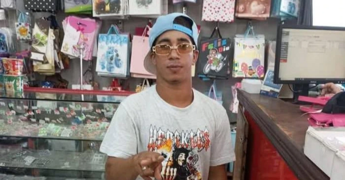 Man in a gray graphic T-shirt and blue backward cap points at the camera in a gift shop at a display-filled counter area.