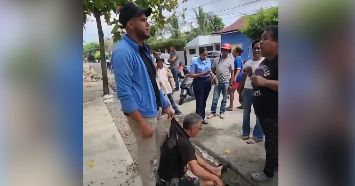 Man in a blue jacket and cap stands with a man sitting on the curb as a crowd watches on a residential street.
