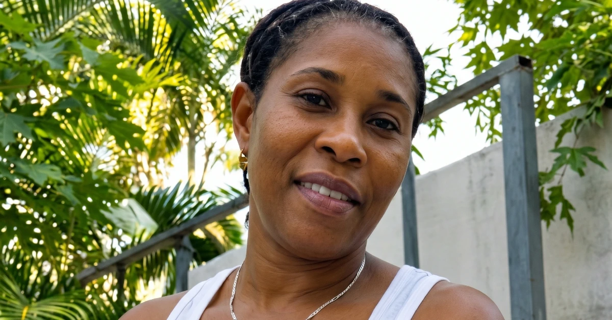 Close-up of a woman smiling outdoors with lush green foliage and a gray fence in the background.
