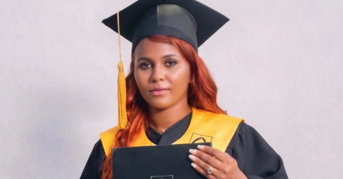 Graduate woman in a black cap and gown with a yellow stole, holding a diploma toward the camera.