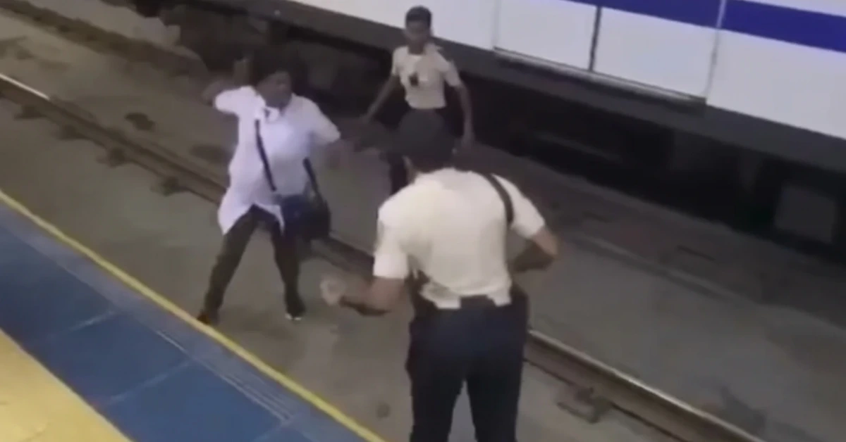 Three people on a train platform; a man in a white shirt raises his arm toward another man while a bystander watches in the background.”,
