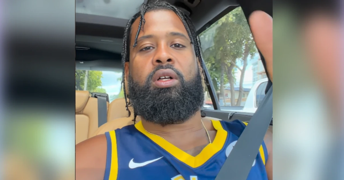 Man with braided hair and beard wearing a blue and gold basketball jersey speaks to the camera from inside a car, seatbelt on.