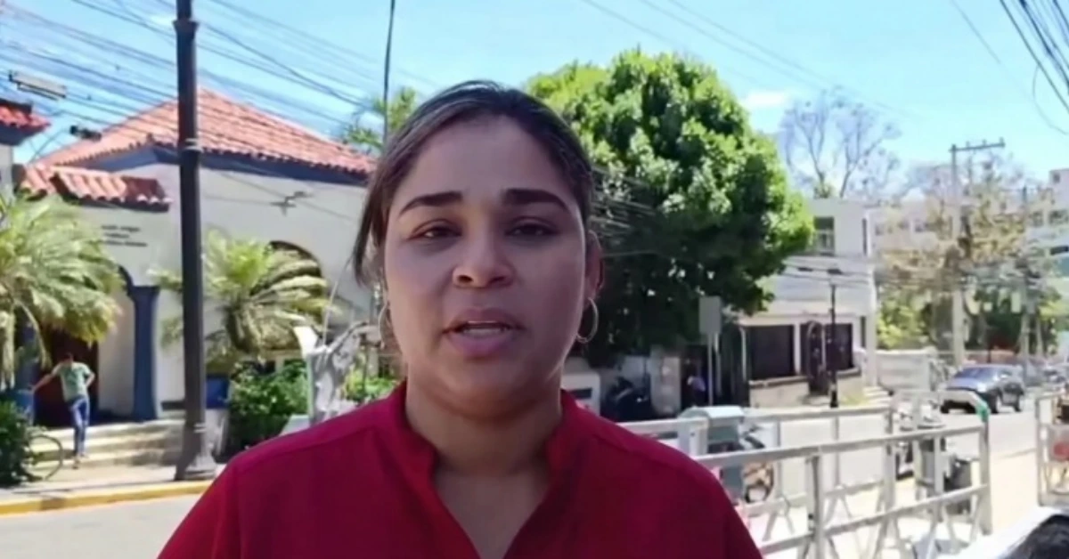 Woman speaks to camera on a sunny street, wearing a red shirt, residential area in the background.