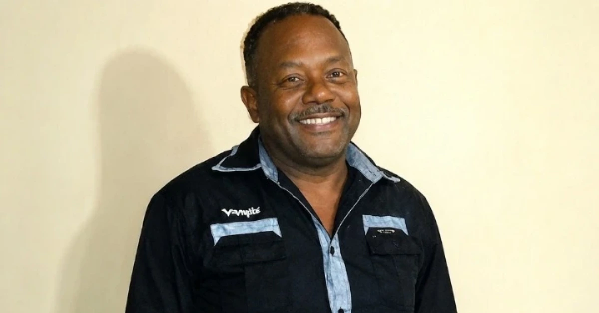 Smiling man in a dark shirt with light blue trim posing against a beige wall.