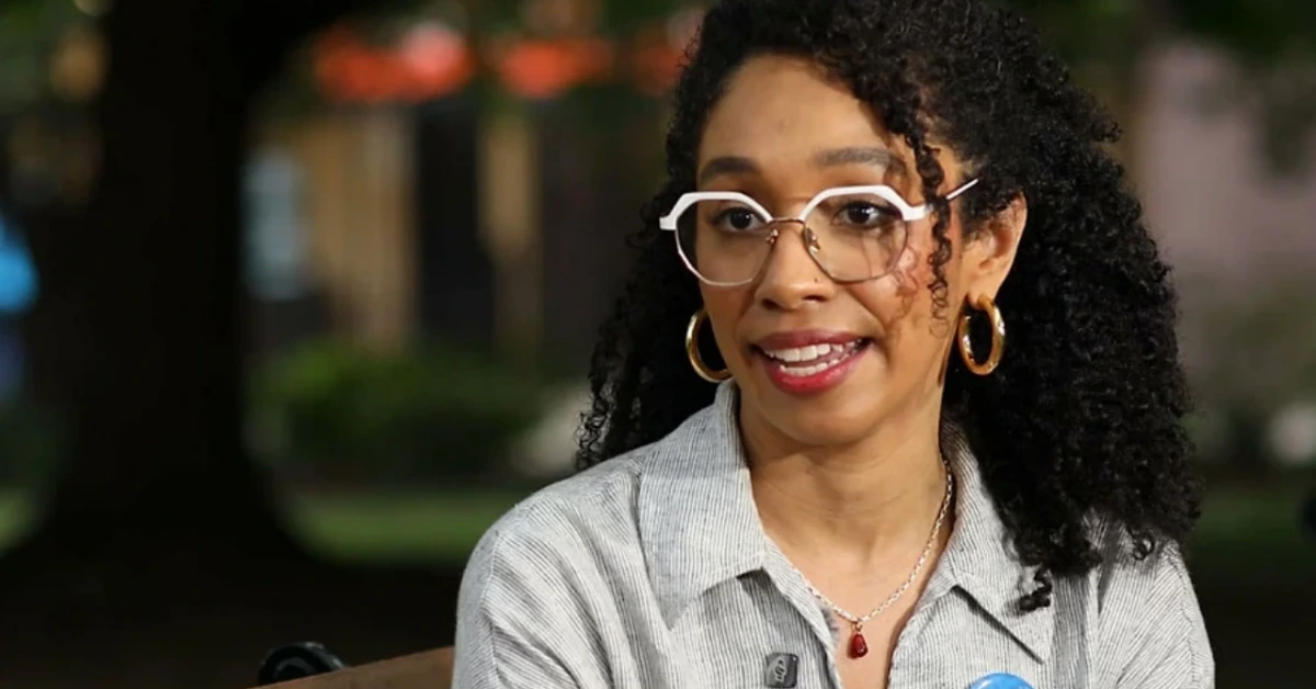 Portrait of a smiling woman with large round white-r framed glasses and gold hoop earrings, outdoors.