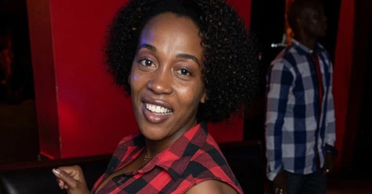 Smiling woman with short curly hair in a red plaid shirt, in a red-lit room; man in a plaid shirt in the background.