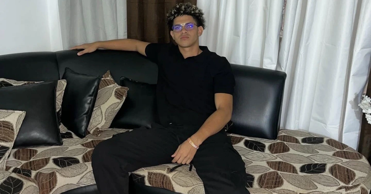Person wearing a black polo and black pants sits casually on a patterned sofa with one arm resting on the back as curtains loom in the background.”,