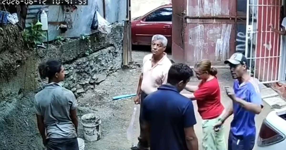 Five people stand in a narrow alley by a stone wall and gate, appearing to discuss or prepare for a small outdoor repair.