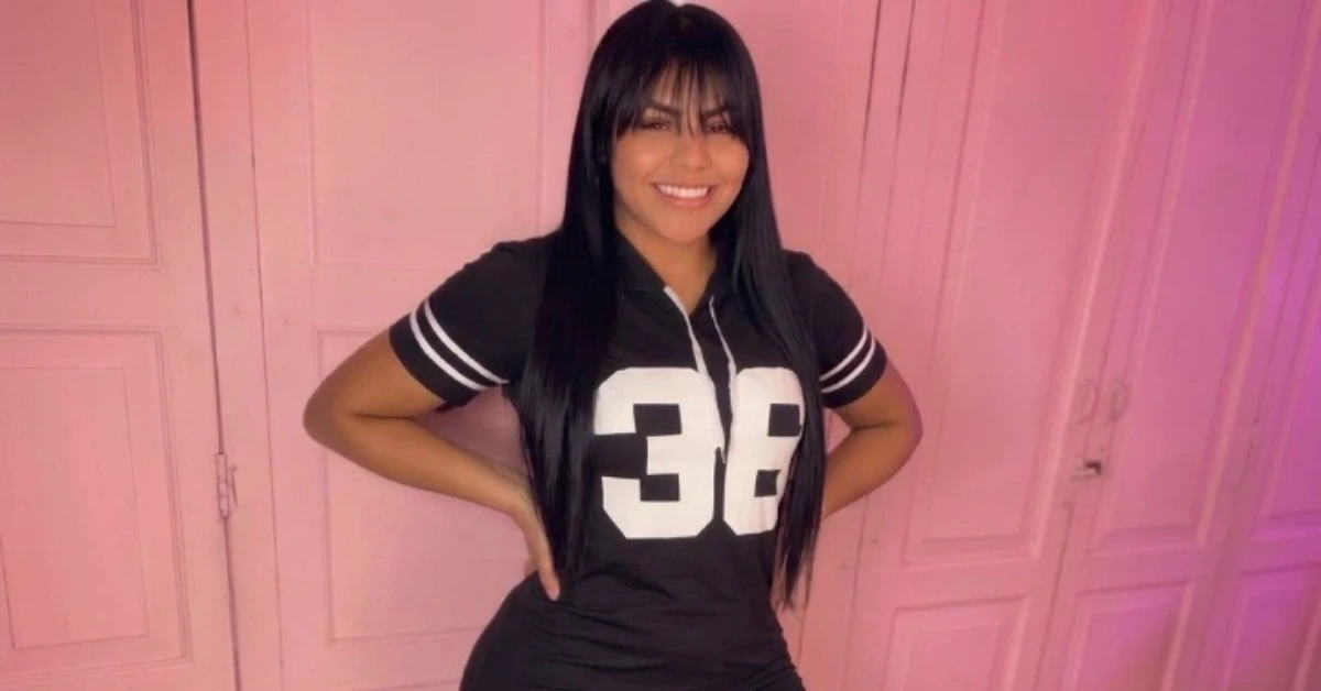 Smiling woman in a black jersey with the number 38, hands on hips, against a pink wall.