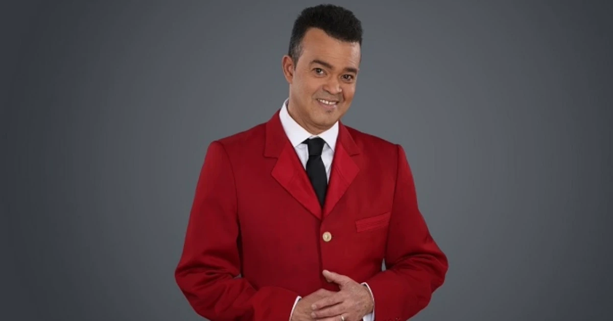 Man in a red blazer, white shirt, and black tie smiles at the camera against a gray backdrop.