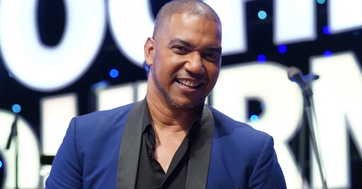 Smiling man in a blue blazer on stage with large lit letters behind him.