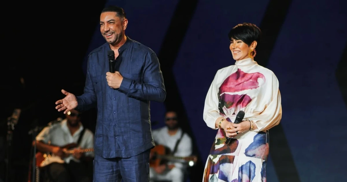 Two hosts on stage, smiling and speaking into wireless microphones, with musicians in the background.