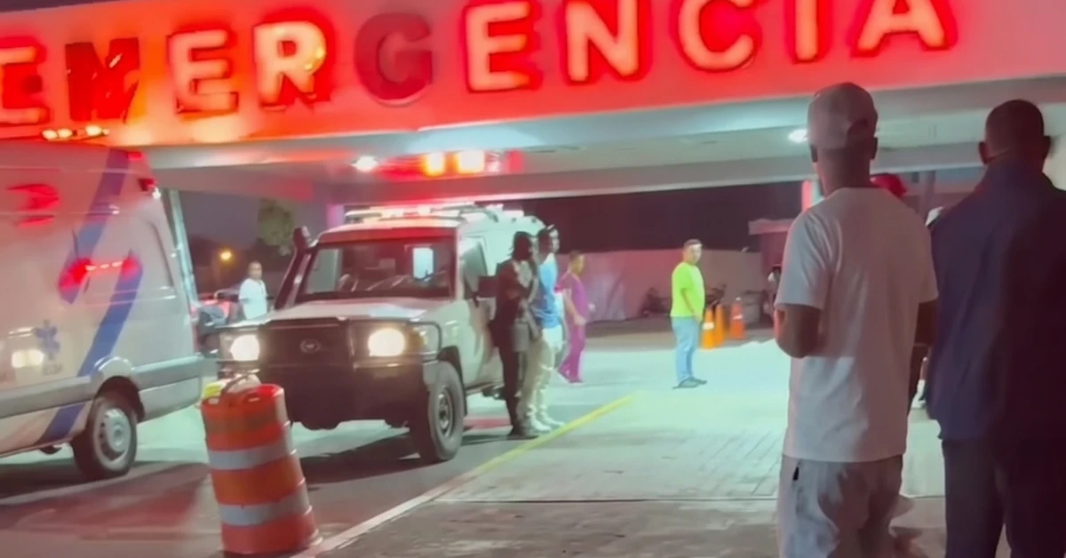 Nighttime hospital emergency entrance with a red neon 'EMERGENCIA' sign, an ambulance and a white van, and people waiting nearby.