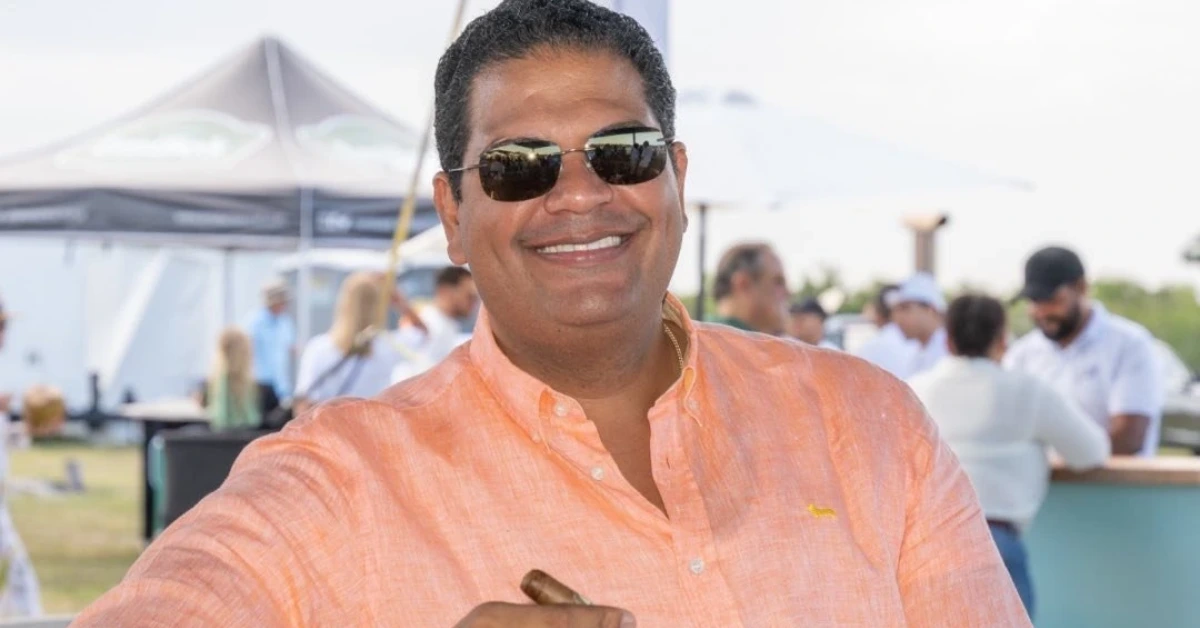 Smiling man in an orange shirt and sunglasses at an outdoor social event with tents in the background.