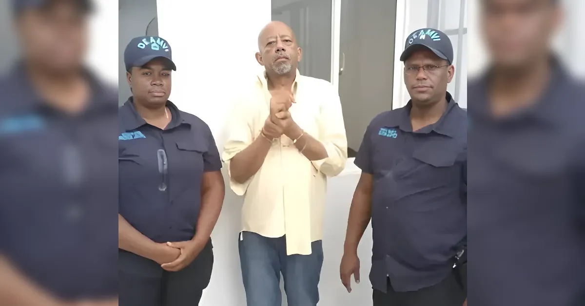 Man in a pale yellow shirt stands center with hands clasped, flanked by two security guards in dark uniforms outside a building.