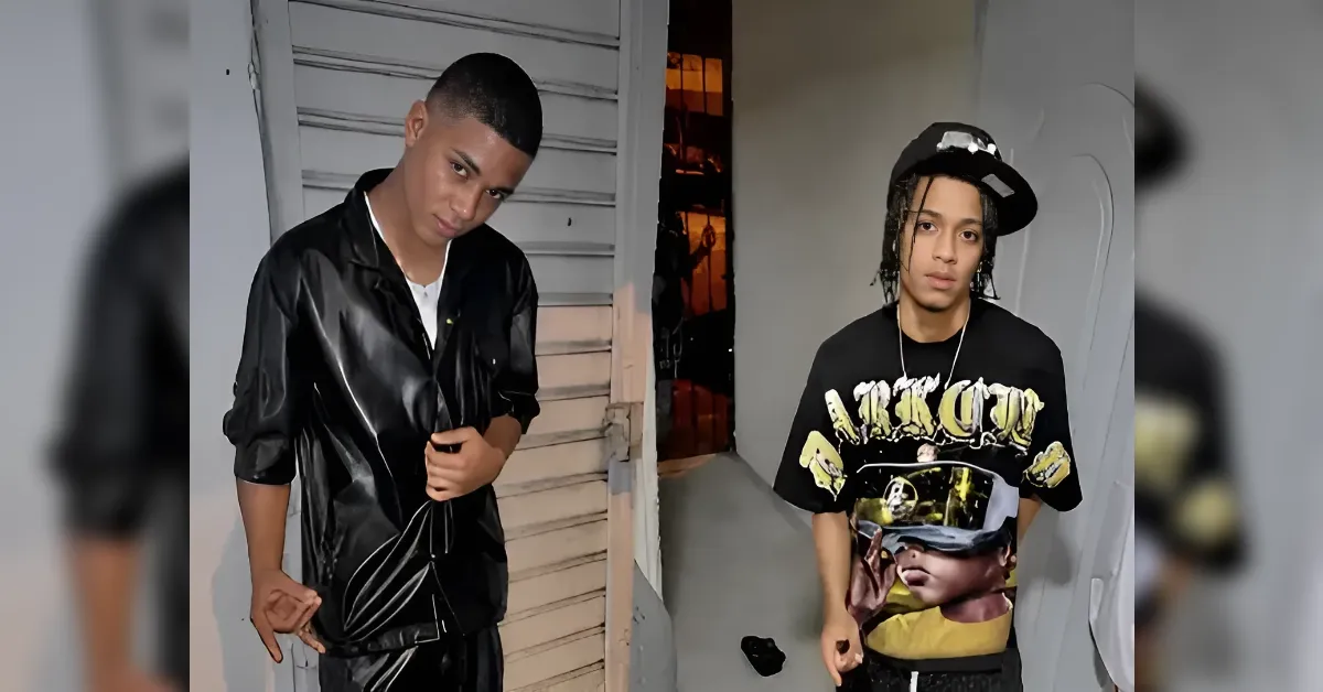 Two young men pose in a doorway hallway; left is in a black shiny jacket over a white shirt, right in a oversized graphic tee and cap with dreadlocks.